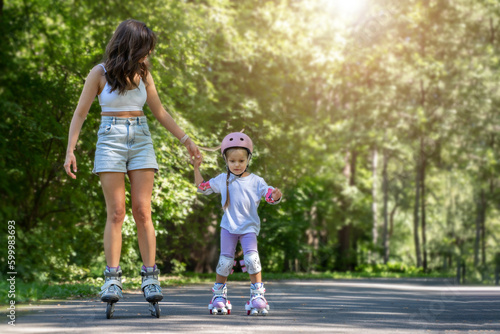 Mother teaching child daughter to skate on inline skates rollers in public park in summer. Family leisure outdoor sport activity game. Copy negative space