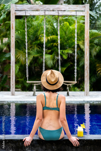 Rear view of woman in bikini sitting on swimming pool ledge relaxing looking at tropical view during vacation. Vertical © Daniel