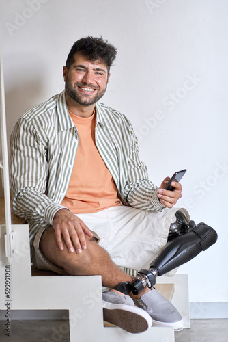 Happy young man with disability above knee transfemoral leg prosthesis sitting on stairs at home holding cell using mobile phone. People with amputation disabilities everyday life, vertical shot. photo