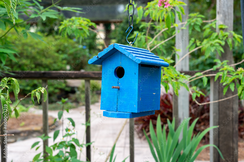 A decorative bright blue single hole birdhouse on a fence. The birdhouse is attached to a wooden deck rail with green trees and shrubs in the background. There's a peg below the small hole. 