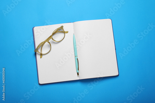Open notebook, pen and glasses on light blue background, top view