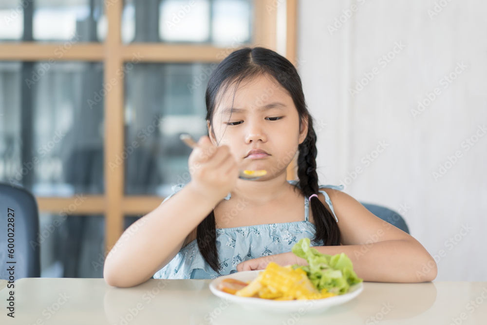 Asian child or kid girl unhappy and don't like eating vegetables salad with corn tomato and greens vegetable with holding spoon or people frowning anorexic bored food at kitchen school or restaurant Asian child or kid girl unhappy and don't like eating vegetables salad with corn tomato and greens vegetable with holding spoon or people frowning anorexic bored food at kitchen school or restaurant