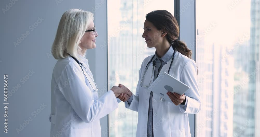 Happy senior and young women physicians shake hands in clinic office, younger thanking for help in teamwork to older colleague, handshaking express gratitude, showing respect in medical collaboration Happy senior and young women physicians shake hands in clinic office, younger thanking for help in teamwork to older colleague, handshaking express gratitude, showing respect in medical collaboration