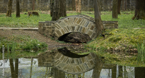 Kamienny mostek pośród drzew z widocznym odbiciem z wodzie. Wokół budząca się do życia zieleń.