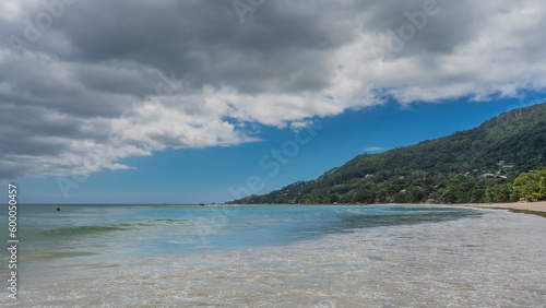 The waves of the turquoise ocean spread over the sandy beach. A hill overgrown with tropical vegetation  against a background of blue sky and clouds. Seychelles. Mahe. Beau Vallon