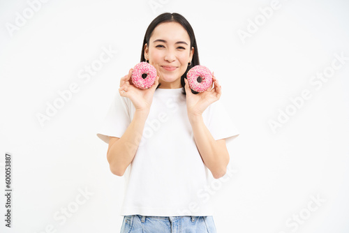 Smiling korean woman eats pink glazed doughnuts, enjoys delicious donnut, junk food, not care about diet, white background photo