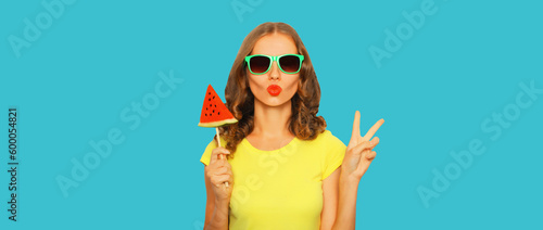 Summer portrait of stylish young woman with fresh juicy lollipop or ice cream shaped slice of watermelon wearing sunglasses on blue background