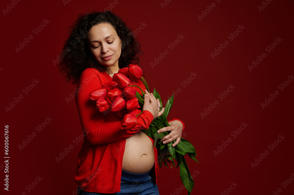 Delightful pretty expectant mother, pregnant woman with a bunch of red tulips, standing bare belly on red color background. Women's and Mother's Day concept. Happy carefree pregnancy and maternity. Delightful pretty expectant mother, pregnant woman with a bunch of red tulips, standing bare belly on red color background. Women's and Mother's Day concept. Happy carefree pregnancy and maternity.