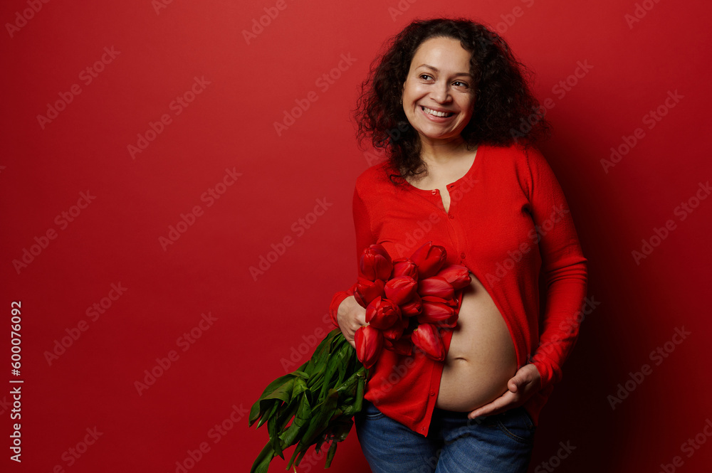 Delightful ethnic pregnant woman holding a bunch of red tulips, touching her belly, mysteriously smiles looking aside over isolated red background. Happy gravid female in 2d semester of her pregnancy Delightful ethnic pregnant woman holding a bunch of red tulips, touching her belly, mysteriously smiles looking aside over isolated red background. Happy gravid female in 2d semester of her pregnancy