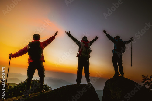 Group of happy hikers or tourists standing with their arms outstretched on the top of the mountain and looking at the sunset.