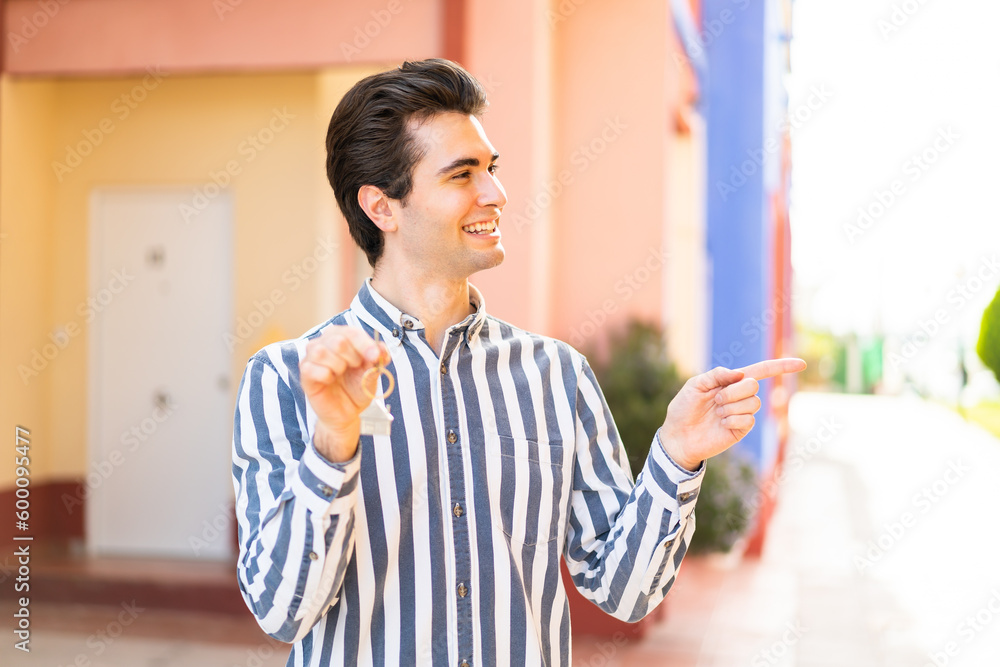 Obraz premium Young handsome man holding home keys at outdoors pointing to the side to present a product