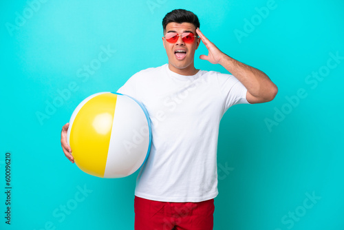Young caucasian man holding a beach ball isolated on blue background with surprise expression
