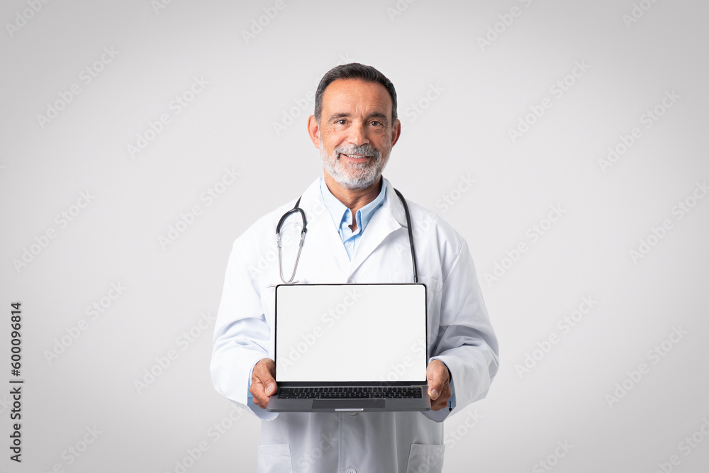 Happy european senior man doctor in white coat with stethoscope shows laptop with blank screen Happy european senior man doctor in white coat with stethoscope shows laptop with blank screen