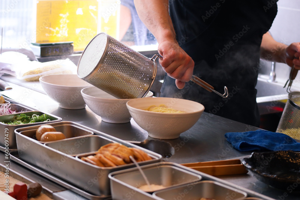 Bowl of Japanese ramen with hands putting the noodles inside it with ramen boiler on a