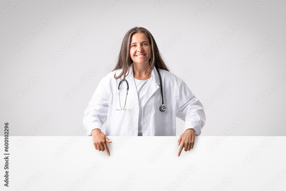 Great medical offer. Happy senior doctor woman pointing down at blank advertising board with empty space for design Great medical offer. Happy senior doctor woman pointing down at blank advertising board with empty space for design