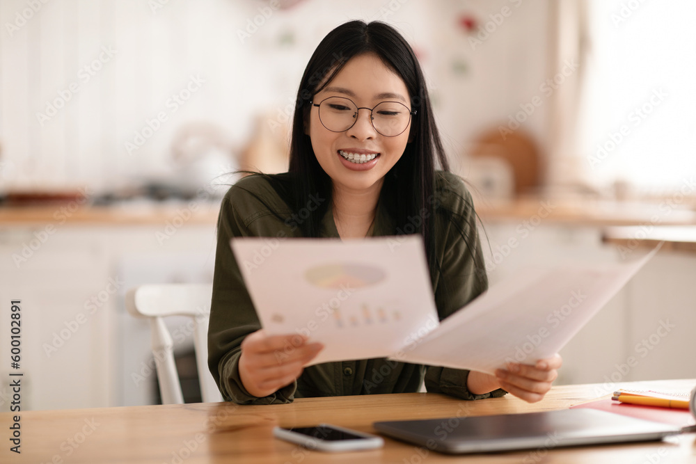 Cheerful young asian woman sitting at kitchen desk, holding papers Cheerful young asian woman sitting at kitchen desk, holding papers