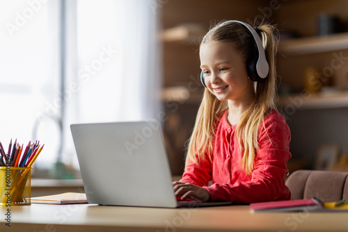 Web-Based Education. Cute Little Schoolgirl Wearing Headphones Study With Laptop At Home