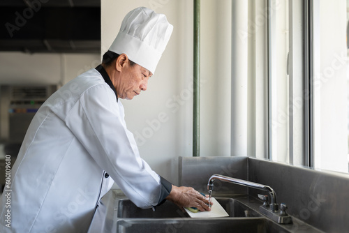 Senior Asian chef man in dress uniform standing washing and cleaning dishes in kitchen restaurant. Cooking and food concept