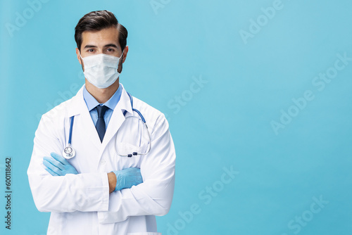 A male doctor in a white coat and a medical mask stands with his hands and looks at the camera on a blue isolated background, copy space, space for text