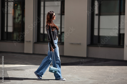Full length beautiful young brunette woman Wearing black leather jacket and wide blue jeans, sneakers, sunglasses and small handbag with chain, walking street on sunny day
