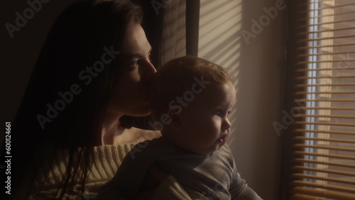 The child in the arms of the mother looks out the window. Millennial mother holds her son in her arms by the window of the house. Blinds on the window. The child's face in the sun. Warm yellow tone.