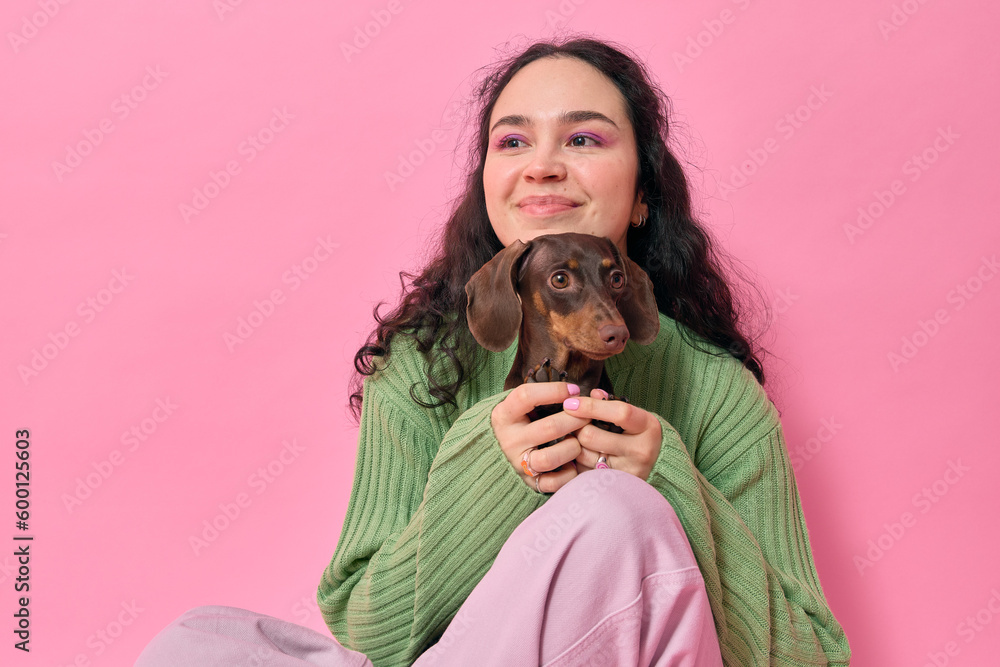 Portrait of young woman with curly hair holding her small brown dachshund puppy on her knees whilst touching his cute little paws smiling looking away and sitting on floor over pink studio background. Portrait of young woman with curly hair holding her small brown dachshund puppy on her knees whilst touching his cute little paws smiling looking away and sitting on floor over pink studio background.