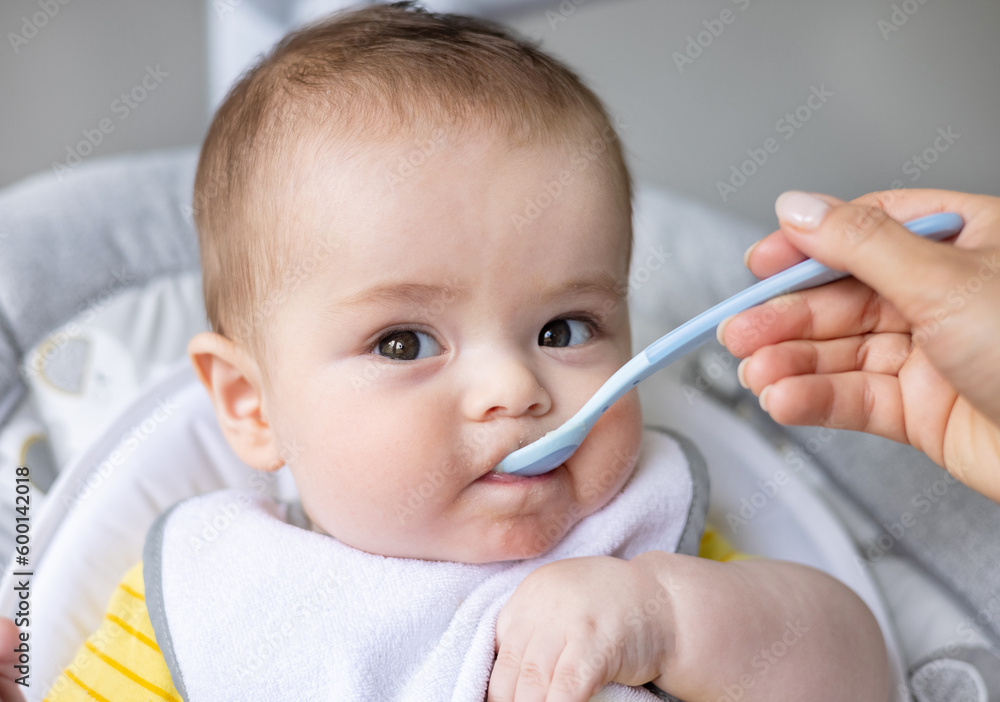 adorable cute baby boy sitting in swing cradle eating mashed food with spoon.mother hand feeding infant toddler.child with bib is smiling,no teeth on gum.vegetables or fruits puree in jar.hungry adorable cute baby boy sitting in swing cradle eating mashed food with spoon.mother hand feeding infant toddler.child with bib is smiling,no teeth on gum.vegetables or fruits puree in jar.hungry