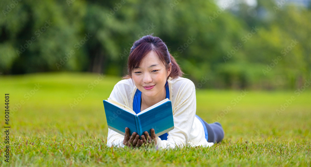 Asian woman is lying down in the grass lawn inside the public park holding book in her hand during summer for reading and education concept