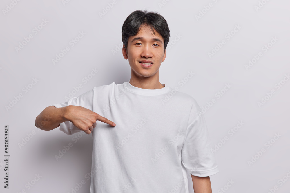 Clothing and design concept. Cheerful dark haired Asian man points at blank space of t shirt suggests to place your promo designing content or advertisement here isolated over white background Clothing and design concept. Cheerful dark haired Asian man points at blank space of t shirt suggests to place your promo designing content or advertisement here isolated over white background