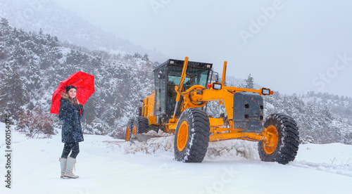 The bulldozer cleans snow on the road, with snowstorm in the background - Beautiful girl in black coat with red umbrella walking down the road while snowfall