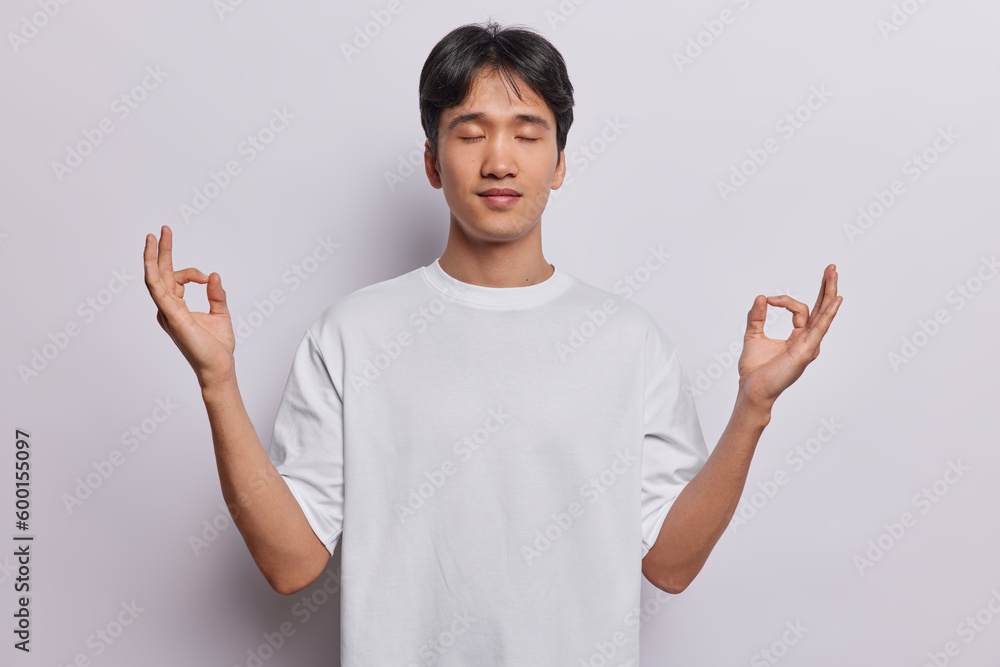 Handsome Chinese man with peaceful expression meditates indoor tries to calm down and relax on break practices yoga dressed in casual t shirt isolated on white background enjoys silence finds balance Handsome Chinese man with peaceful expression meditates indoor tries to calm down and relax on break practices yoga dressed in casual t shirt isolated on white background enjoys silence finds balance