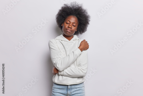 Indoor shot of young Afro American woman embraces herself with love crosses arms over body recalls pleasant moment dressed in comfortable sweatshirt and jeans isolated over white background.