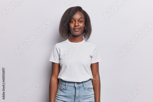 Studio shot of attractive woman with Afro hair has dark healthy skin looks calmly at camera poses in studio wears casual t shirt and jeans isolated over white wall. Dark skinned female model indoors photo