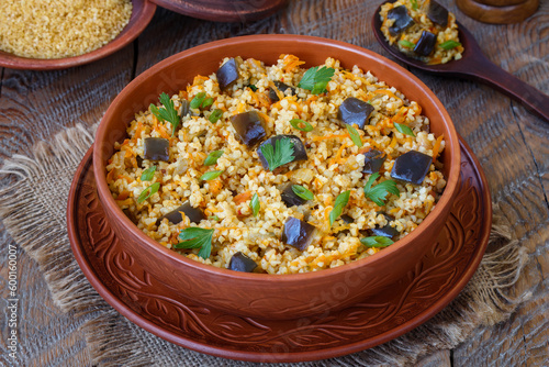 Vegetable Bulgur Pilaf. Spiced Bulgur with eggplant, onion, carrot and herbs served in a clay bowl on wooden table. Selective focus, horizontal.