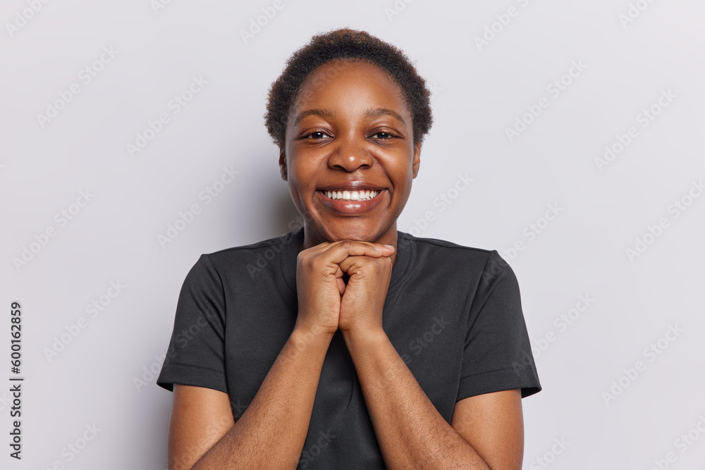 Portrait of cheerful dark skinned woman with short hair keeps hands under chin smiles toothily dressed in casual black t shirt expesses sincere positive emotions isolated over white background Portrait of cheerful dark skinned woman with short hair keeps hands under chin smiles toothily dressed in casual black t shirt expesses sincere positive emotions isolated over white background