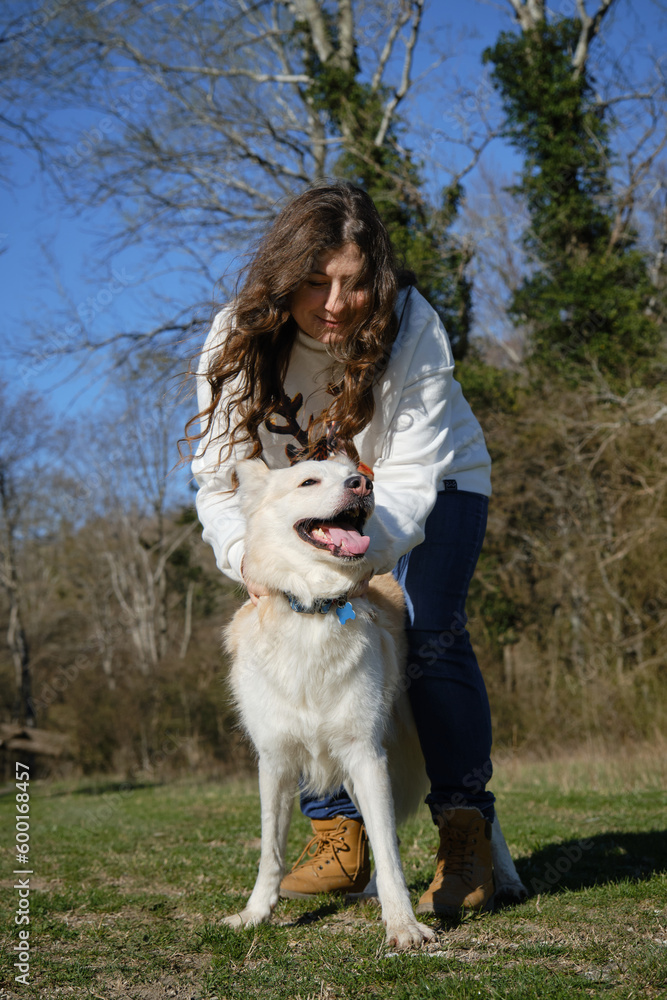 Caucasian woman walks in field with white dog of mixed breed. Female owner has fun with pet outside in clearing near forest in spring. Girl with wavy hair strokes and hugs half breed Swiss Shepherd. Caucasian woman walks in field with white dog of mixed breed. Female owner has fun with pet outside in clearing near forest in spring. Girl with wavy hair strokes and hugs half breed Swiss Shepherd.