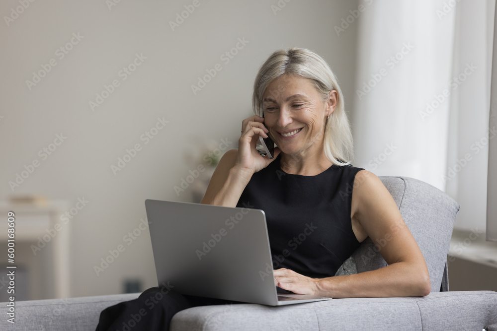 Happy positive blonde mature professional woman talking on mobile phone at laptop at home, enjoying online wireless communication, connection, resting in armchair, smiling, laughing Happy positive blonde mature professional woman talking on mobile phone at laptop at home, enjoying online wireless communication, connection, resting in armchair, smiling, laughing