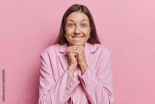 Portrait of dark haired woman bites lips keeps hands under chin looks directly at camera dressed in formal clothes isolated over pink background works in business sphere anticipates for positive news
