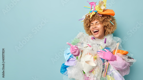 Overjoyed woman covered with plastic rubbish wears protective gloves points finger aside on blank space shows something funny collects garbage to clean out our planet stands against blue background