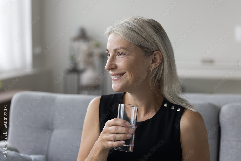 Cheerful happy pretty mature lady holding glass of natural mineral water, sitting on couch at home, looking away with toothy smile, thinking over hydration, metabolism, healthcare Cheerful happy pretty mature lady holding glass of natural mineral water, sitting on couch at home, looking away with toothy smile, thinking over hydration, metabolism, healthcare