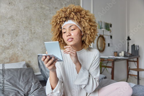 Serious attractive curly haired woman applies anti fatigue undereye mask concentrated in mirror touches chin gently dressed in domestic clothes poses on bed against cozy interior. Beauty treatments photo