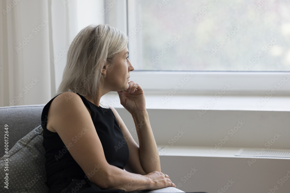 Thoughtful depressed mature woman sitting on couch at home, looking at window away in deep thoughts, suffering from depression, apathy, emotional crisis, thinking over problem, loss - obrazy, fototapety, plakaty 