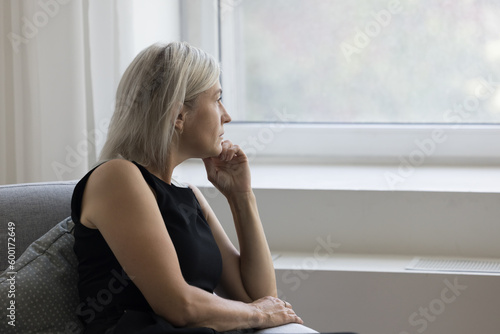 Fototapeta Thoughtful depressed mature woman sitting on couch at home, looking at window aw