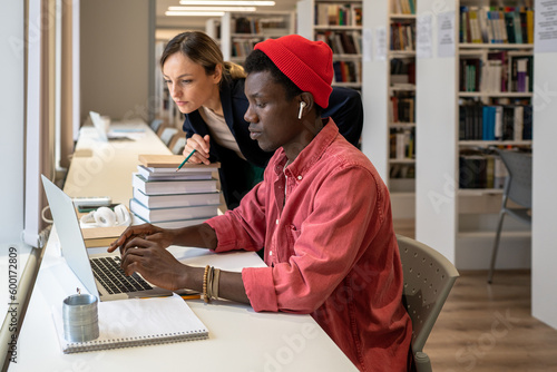 Interested concentrated african american student man typing on laptop near serious college tutor woman looking at screen holding pencil. Focused diverse couple working on project prepare to exam test
