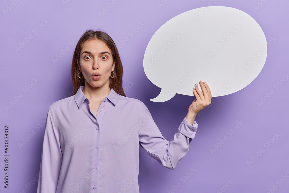 Stunned young female office worker dressed in shirt holds empty speech bubble for your information or advertisement isolated on purple background. Surprised European woman suggests to place your idea Stunned young female office worker dressed in shirt holds empty speech bubble for your information or advertisement isolated on purple background. Surprised European woman suggests to place your idea