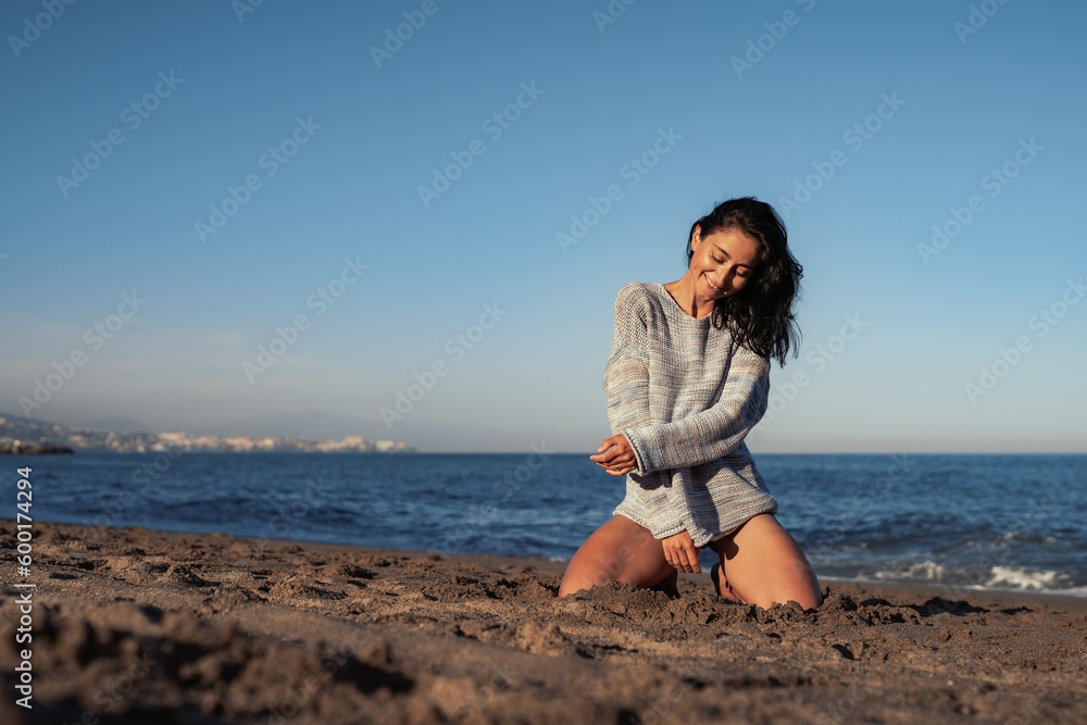 beautiful brunette woman in a sweater and swimsuit sits on the sand by the sea enjoys the environment