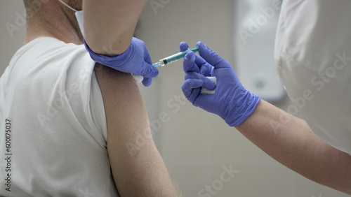 A female doctor vaccinates a young guy. Vaccination against coronavirus. Girl doctor holds a syringe in her hands, inserts a needle into the arm muscle photo