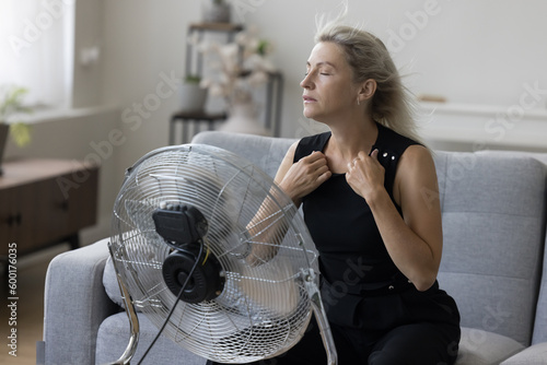 Sick exhausted senior woman with flying blonde hair and closed eyes hair sitting on couch at fresh air blowing from propeller, getting cool, refreshed at fan, feeling overheated, stress relief photo