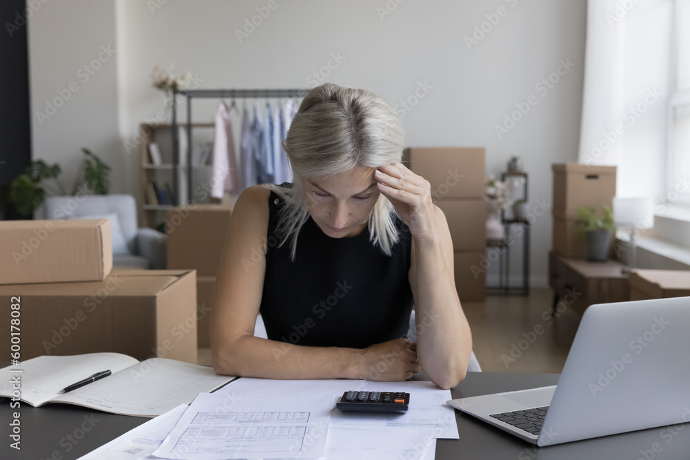Middle aged entrepreneur about to bankrupt. Mature small business owner woman staring at paper invoices, calculator, holding head, feeling depressed, frustrated, desperate Middle aged entrepreneur about to bankrupt. Mature small business owner woman staring at paper invoices, calculator, holding head, feeling depressed, frustrated, desperate