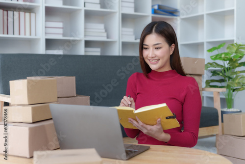 Smiling young Asian business owner woman prepare parcel box and check online orders of product for deliver to customer on laptop computer at home. Shopping Online concept. Startup small business SME.
